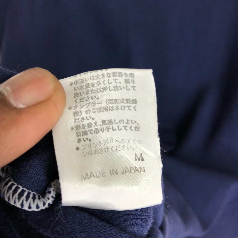 May include: White tag with black text detailing care instructions for a navy blue shirt. The tag includes the size "M" and the words "Made in Japan".