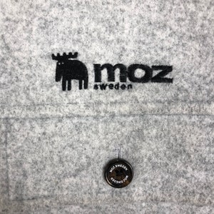 May include: A gray felt fabric with a black embroidered moose and the text "moz sweden". A black button with the text "moz sweden" is also visible.