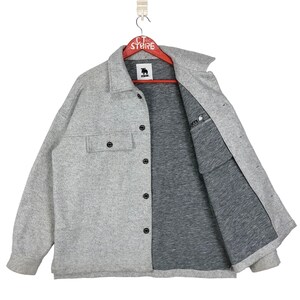 May include: A gray long-sleeved shirt with a button-up front and a single chest pocket. The shirt has a black logo on the left side of the chest that says "MUCK".