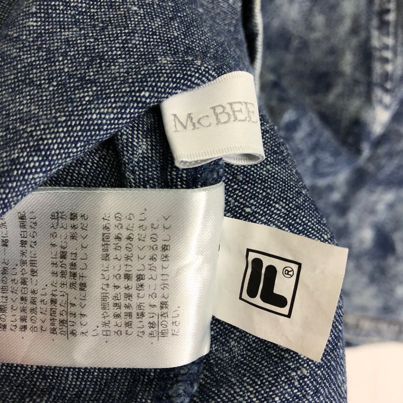 Op de afbeelding: Een wit label met de tekst "McBEE" in zwarte letters. Een wit label met de tekst "IL" in zwarte letters. De labels zijn bevestigd aan een blauwe denim stof met een wit splatterpatroon.