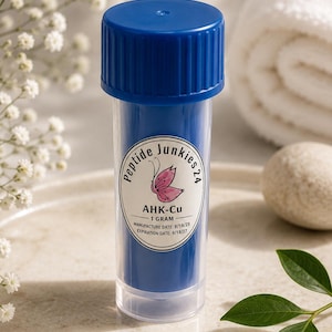 May include: A small, clear plastic vial with a blue screw-top lid containing blue powder. The label reads Peptide Junkies 24, AHK-Cu, 1 gram, with a manufacture date of 18/9/25 and an expiration date of 18/9/27, featuring a pink butterfly icon.