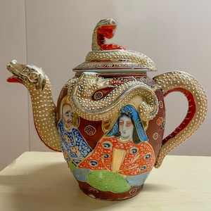 May include: A decorative ceramic teapot with a dragon-shaped spout and handle. The teapot is primarily maroon with gold and white accents. It features painted figures and a snake-like design on the lid. The teapot is a unique piece of Asian-inspired decor.