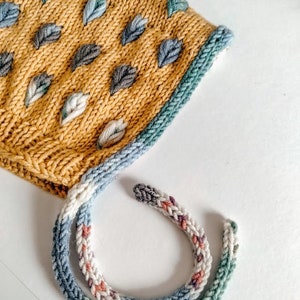 Può includere: Un berretto per neonato lavorato a maglia con una base gialla e un motivo a foglie blu, verde e bianco. Il berretto ha un lungo laccio multicolore.