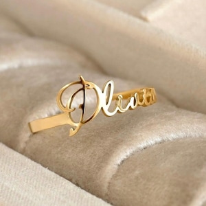 Peut inclure: Une bague en métal doré portant le prénom Olivia écrit dans une police cursive. Les lettres sont reliées à un anneau fin et poli qui entoure le doigt. Le design présente une finition métallique brillante avec une texture lisse.
