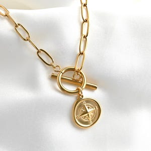 May include: Gold-coloured necklace with a compass pendant. The necklace has a toggle clasp and a chain with elongated oval links. The compass pendant is round with a detailed design, including directional markers. The necklace is displayed on a white, silky background.