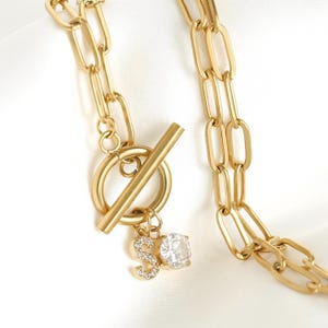 Peut inclure: Un collier à chaîne doré composé de maillons ovales allongés et d'un fermoir à bascule. Une petite breloque en forme de lettre S, incrustée de cristaux transparents, est suspendue à l'anneau du fermoir avec un cristal rond transparent.
