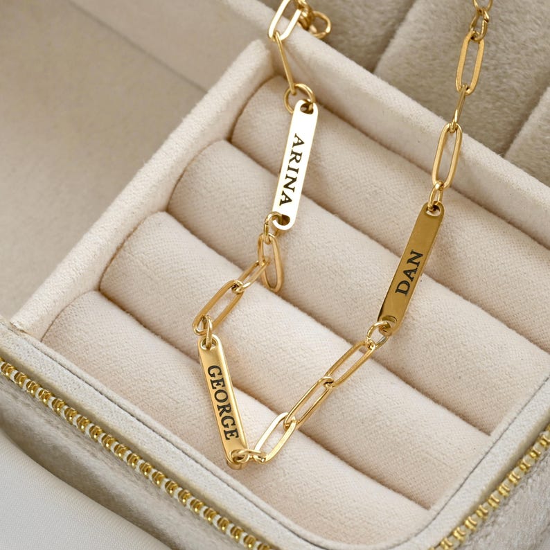 May include: A gold-toned chain necklace featuring elongated oval links interspersed with three rectangular metal bars. Each bar is engraved with a name in black capital letters: ARINA, DAN, and GEORGE, integrated directly into the chain structure.