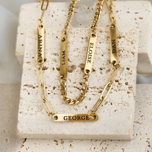 Può includere: Una collana a strati color oro composta da diverse catene con barrette metalliche rettangolari. Ogni barretta è incisa con un nome: Arina, Liam, Eloise, Dan e George. Il design combina catene a maglie sottili con una catena a maglia gourmet.