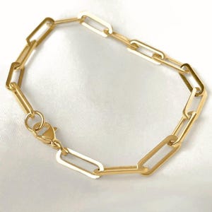 May include: A gold-toned chain bracelet featuring elongated, rectangular paperclip-style links. The bracelet is secured with a lobster claw clasp and a small circular jump ring, showcasing a polished, metallic finish throughout the entire piece.