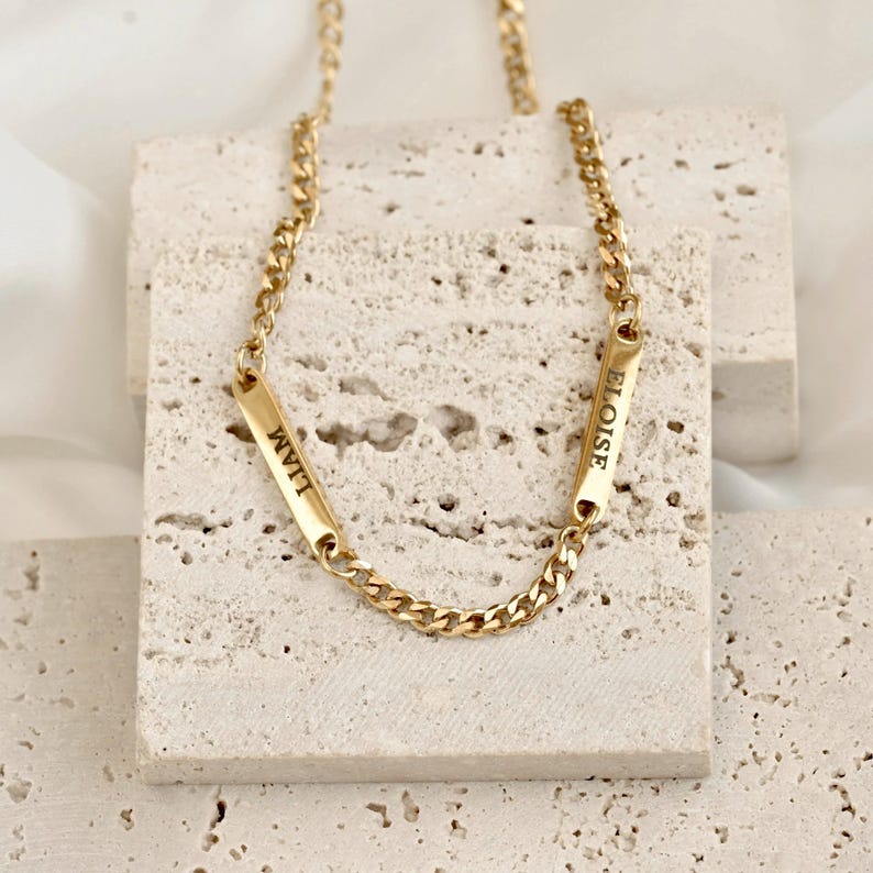 May include: A gold-toned chain necklace featuring two rectangular metal bars integrated into the links. Each bar is engraved with a name in black capital letters, with one bar reading LIAM and the other bar reading ELOISE in a clear font.