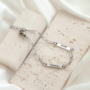 May include: A silver-toned metal chain bracelet featuring two rectangular bar charms. One bar is engraved with the name Jamie and the other with the name Jessica. The bracelet includes a lobster claw clasp and a short length of extender chain.