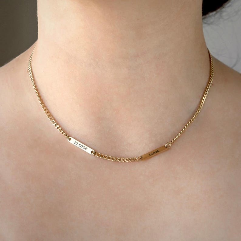 May include: A gold-toned chain necklace featuring two small, rectangular metal bars integrated into the chain. Each bar is engraved with a name, with the left bar reading ELOISE and the right bar reading LIAM in a clean, sans-serif font style.