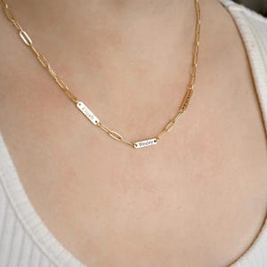 May include: A gold-toned chain necklace featuring elongated oval links and three small, rectangular metal tags integrated into the chain. Each tag is engraved with a name: Elijah, Wesley, and Parker, in a clear, dark, sans-serif font.