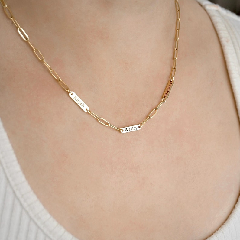 May include: A gold-toned chain necklace featuring elongated oval links and three small, rectangular metal tags integrated into the chain. Each tag is engraved with a name: Elijah, Wesley, and Parker, in a clear, dark, sans-serif font.