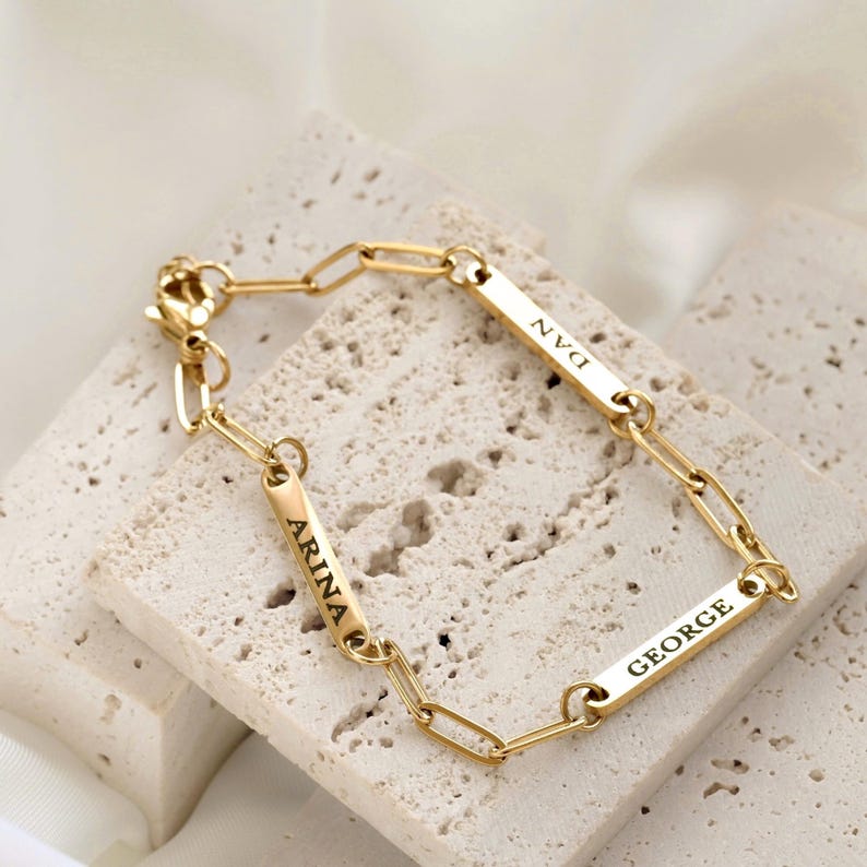 May include: A gold-toned chain link bracelet featuring three rectangular metal bars, each engraved with a name. The names ARINA, DAN, and GEORGE are clearly visible in black lettering on the polished gold surfaces of the individual bars.