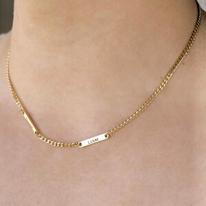 May include: A gold-toned chain necklace featuring two small, rectangular metal bars integrated into the chain. One bar is engraved with the name LIAM, while the other bar is engraved with a name that appears to be ELOISE in a vertical orientation.