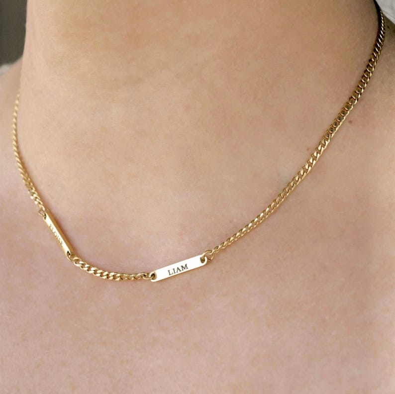 May include: A gold-toned chain necklace featuring two small, rectangular metal bars integrated into the chain. One bar is engraved with the name LIAM, while the other bar is engraved with a name that appears to be ELOISE in a vertical orientation.