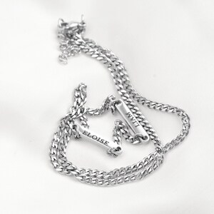 May include: A silver-toned metal chain bracelet featuring two rectangular bar charms. Each bar is engraved with a name, one reading ELOISE and the other reading LIAM. The bracelet has a classic curb chain link design and a small lobster claw clasp.