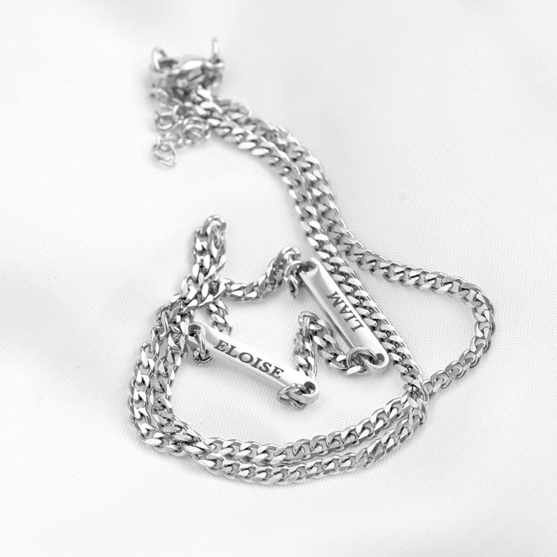 May include: A silver-toned metal chain bracelet featuring two rectangular bar charms. Each bar is engraved with a name, one reading ELOISE and the other reading LIAM. The bracelet has a classic curb chain link design and a small lobster claw clasp.
