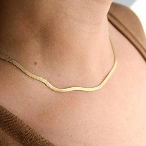 May include: A gold-toned herringbone chain necklace featuring a flat, flexible, and tightly woven link design that rests against the neck. The metal surface has a polished, reflective finish that catches the light, creating a smooth, continuous appearance.