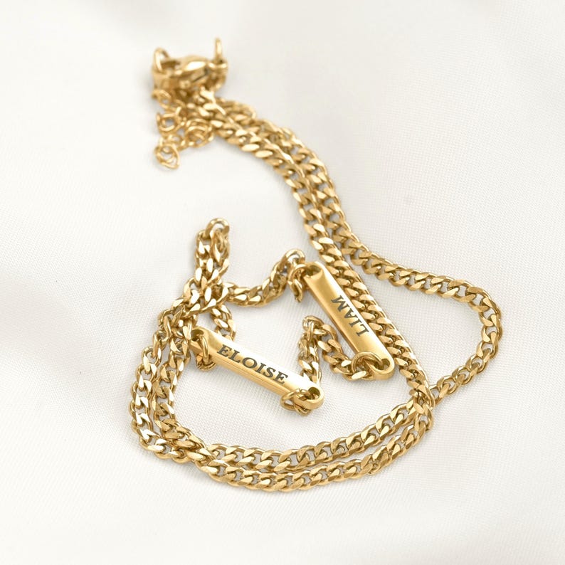May include: A gold-toned metal chain bracelet featuring two rectangular bar charms. One bar is engraved with the name ELOISE and the other bar is engraved with the name LIAM. The bracelet includes a lobster claw clasp and an adjustable extender chain.