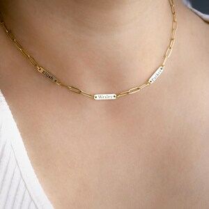 May include: A gold-toned chain necklace featuring three small, rectangular metal plaques integrated into the links. Each plaque is engraved with a name: Elijah, Wesley, and Parker. The chain consists of elongated, oval-shaped links in a uniform design.