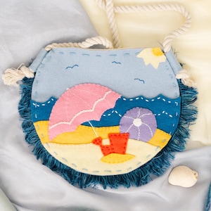 May include: A felt beach-themed purse with a rope strap, featuring a pink umbrella, a purple umbrella, and a red bucket on a yellow beach. A light green shovel, a felt fish, and a felt jellyfish are also present.