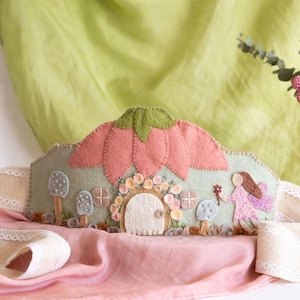 May include: A felt fairy crown with a pink flower design and green leaves. The crown has a light green base with mushroom and fairy details. A pink ribbon with lace trim is draped in front of a green and pink backdrop.