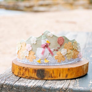 May include: A decorative felt crown with a goose character, pink bow, and balloon. The crown is adorned with felt flowers and lace, presented on a wooden base. A delightful accessory for a child's birthday or special event, measuring approximately 20 cm in diameter.