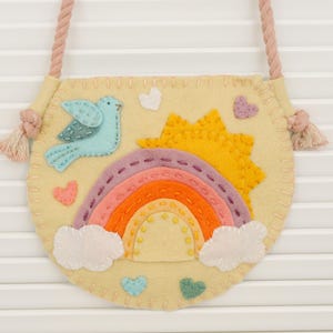 May include: A round, yellow felt bag with a rainbow, sun, bird, and heart appliqués. The bag has a pink rope strap with tassels. The rainbow features orange, pink, and purple arches. The bird is light blue.
