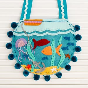 May include: A turquoise felt purse with a whimsical underwater scene. The design includes a jellyfish, fish, seaweed, and an anchor. The purse has a blue pom-pom trim and turquoise straps with a scalloped edge.