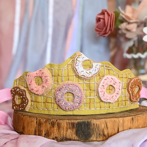 May include: A yellow felt crown decorated with various donut designs in pink, white, and brown. The crown sits on a wooden base with a pink ribbon attached. The background includes flowers and draped fabric.