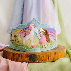 May include: A felt crown with a whimsical design. The crown features a unicorn, a girl in a pink dress, and a rainbow. The crown is set on a wooden base. The background includes pastel fabrics.