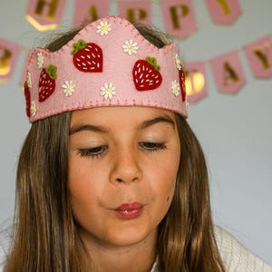 Könnte beinhalten: Ein rosa Filzkrone, verziert mit roten Erdbeeren und weißen Gänseblümchen. Die Krone wird von einer jungen Person getragen. Ein rosa Banner mit goldenen Buchstaben, die "HAPPY DAY" buchstabieren, ist im Hintergrund zu sehen.