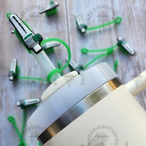 May include: A white and cream colored Stanley brand travel mug with a stainless steel lid and a straw. The mug has a brown bear logo with a crown on its head. The straw is attached to a green silicone strap with a small green and white plastic clip.
