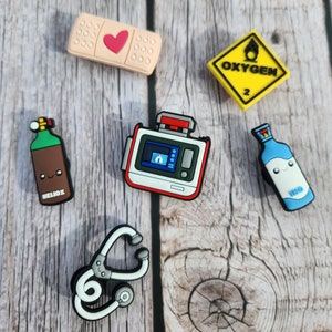 May include: A set of seven colorful medical themed shoe charms. The charms include a bandage with a heart, an oxygen tank, a stethoscope, a bottle of medicine, a medical device, and a sign that says "Oxygen".