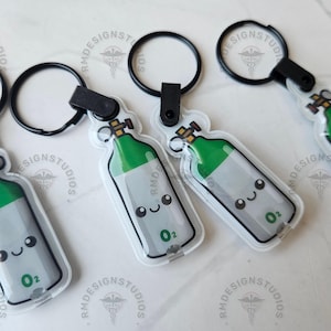 May include: Several oxygen tank keychains with black key rings. The keychains are clear with a cartoon oxygen tank design in green, gray, and white. Each tank has a smiling face and the letters "O2".