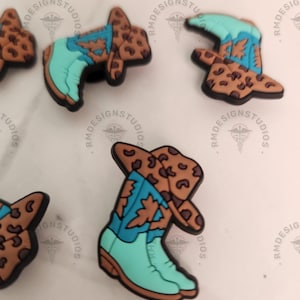 May include: A set of four charm shoe charms featuring a cowboy boot with a cowboy hat on top. The charms are brown and tan with a turquoise blue boot and hat. The charms are made of a soft, flexible material.