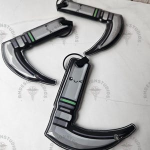 May include: Several gray and black keychains in the shape of a curved blade, with a green stripe and a small smiling face on one. The keychains are arranged on a white surface.