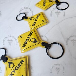 May include: Several yellow keychains with black text that reads "OXYGEN" and the number "2". Each keychain has a black ring attached. The keychains are on a white surface. The keychains are from RM Design Studios.