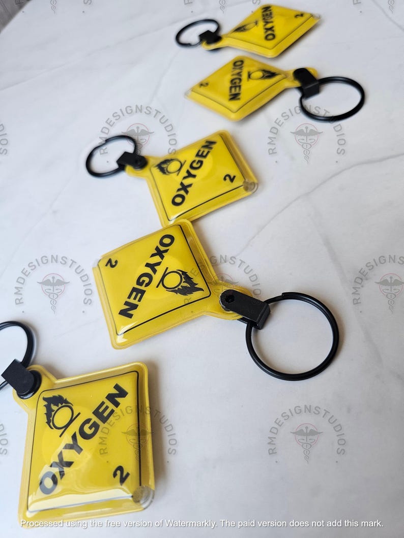 May include: Several yellow keychains with black text that reads "OXYGEN" and the number "2". Each keychain has a black ring attached. The keychains are on a white surface. The keychains are from RM Design Studios.