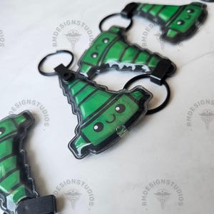 May include: Green crayon-shaped keychains with black accents and a smiling face. The keychains are attached to black key rings and are displayed on a white surface. The image includes the text "RM DESIGN STUDIOS".