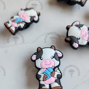 May include: A set of five cow-shaped shoe charms. The charms are made of a soft, flexible material and feature a black and white cow pattern with pink accents. Each charm has a different expression and pose, including a cow holding a blue and pink ice cream cone.