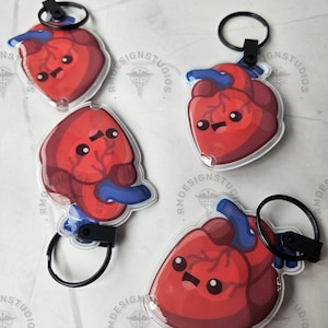 May include: Four red heart-shaped keychains with blue veins and black key rings. Each keychain has a cartoon face. The keychains are on a white surface with the text "RMDESIGNSTUDIOS".