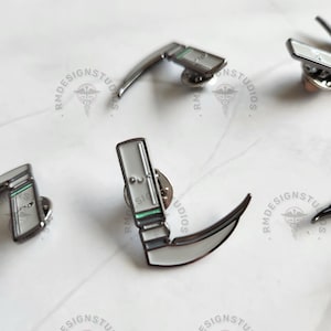May include: A set of five enamel pins featuring a medical instrument, a scalpel, with a green and white handle. The pins are silver with black outlines.