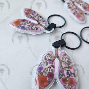 May include: Clear acrylic keychains in the shape of lungs, adorned with a floral design in shades of pink, orange, purple, and green. Each keychain has a black keyring and a black strap. The keychains are scattered on a white surface.