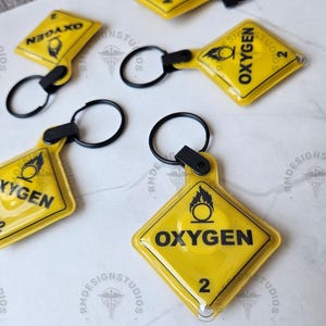 May include: Yellow diamond-shaped keychains with the word "OXYGEN" in black, a flame symbol, and the number "2". Each keychain has a black ring and clasp. The keychains are on a white surface.