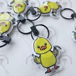 May include: Yellow chick-shaped keychains with black key rings. The cartoon chick has a black outline, orange beak and feet, and is holding a hammer and a blue object. The keychains are on a white surface.