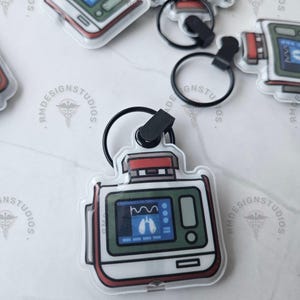 May include: A clear acrylic keychain featuring a medical device design with a screen displaying a lung diagram. The keychain has a red and white border, a black key ring, and a black clasp. The design is a medical theme.