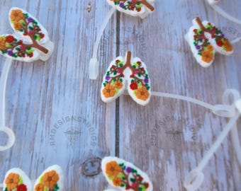 May include: A set of colourful, floral-patterned, silicone lung charms. The charms are attached to clear, plastic lanyards. The charms are a light white colour with a brown tree trunk and colourful flowers.